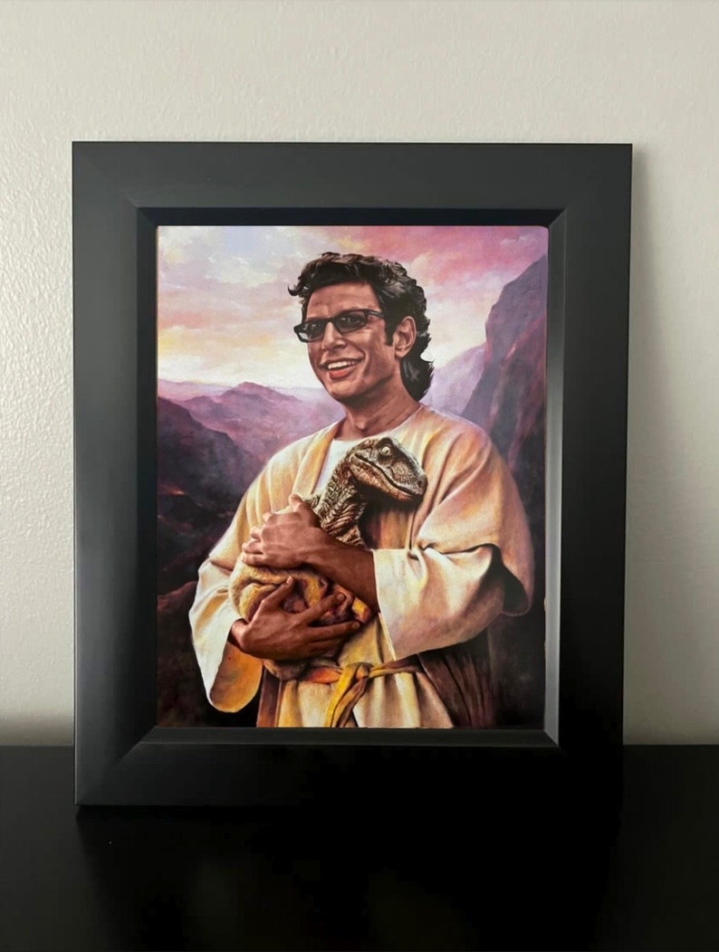 May include: Framed print featuring a portrait of a man in sunglasses holding a small dinosaur. The artwork is set against a backdrop of a mountain landscape with a sunset. The frame is black.