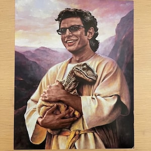 May include: A print featuring a smiling man in glasses holding a baby dinosaur. The man is wearing a cream-colored robe, set against a backdrop of mountains and a sunset sky. The dinosaur is a mix of brown and green.