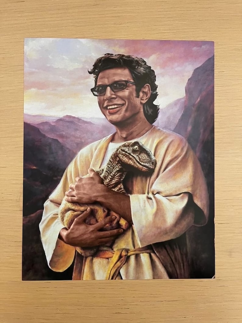 May include: A print featuring a smiling man in glasses holding a baby dinosaur. The man is wearing a cream-colored robe, set against a backdrop of mountains and a sunset sky. The dinosaur is a mix of brown and green.