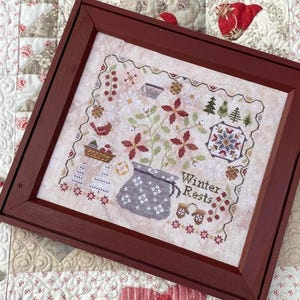 May include: A framed cross-stitch sampler with a winter theme. The artwork features floral and snowflake motifs, a teapot, and the words "Winter Rests." The frame is a dark reddish-brown colour, and the background is a quilted fabric.