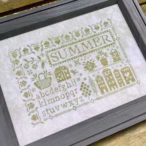 May include: A framed cross-stitch sampler with the word "SUMMER" in olive green. The design includes floral and house motifs, a basket, potted plants, and the alphabet. The frame is a dark gray.