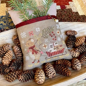 Può includere: Una pochette beige a tema natalizio con un disegno a punto croce di un topo che tira una slitta piena di regali. La pochette è decorata con fiocchi di neve e rametti di verde. La pochette è circondata da pigne e rametti di sempreverdi.