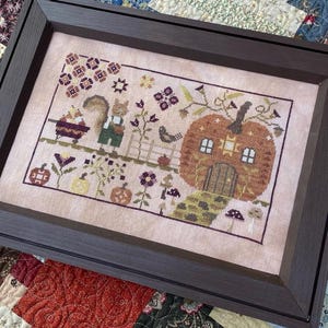 May include: Framed cross-stitch artwork featuring a whimsical autumn scene. A squirrel in overalls stands near a pumpkin house with a door and windows. The design includes flowers, a bird, and a fence, all in warm, earthy tones.
