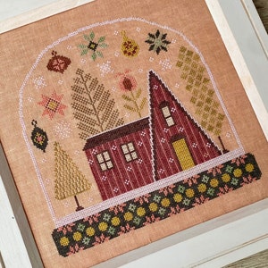 May include: Cross-stitch artwork in a white frame. The design features a red A-frame house and a smaller house, with trees and decorative elements inside a snow globe shape. The background is a peach colour, and the frame is white.