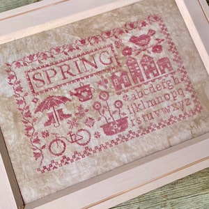 Op de afbeelding: Kruissteekborduurwerk in een lichtroze lijst. Het ontwerp toont het woord "SPRING" en verschillende lentemotieven zoals een vogel, huizen en een fiets, allemaal in rode draad. De achtergrond is een neutrale beige stof.