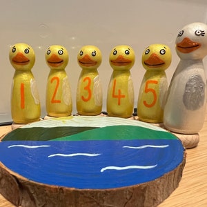 May include: Five yellow wooden ducks with orange numbers 1 to 5 painted on them, standing on a wooden base with a blue lake and green hill painted on it. A white wooden duck with grey wings is standing behind the ducks.