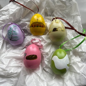 May include: Five wooden Easter egg ornaments featuring different religious themes. The ornaments are painted in various colours, including purple, yellow, green, pink, and white. The themes include a dove, a chalice and bread, a palm branch, a tomb, and a cross.