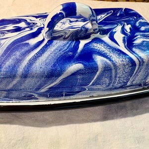 May include: A rectangular butter dish with a lid featuring a glossy blue and white marbled pattern. The lid has a curved handle on top, and the base is a white tray with a thin black rim, designed to hold the lid securely in place for storage.
