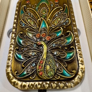 May include: A rectangular metal compact mirror featuring a raised peacock design in antique gold finish with teal enamel accents and embedded multicolored rhinestones in shades of orange, green, and light blue, set within a textured border.