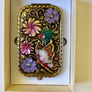 May include: A rectangular metal compact mirror featuring an antique brass finish with embossed floral and butterfly motifs. The design is accented with purple, pink, and teal enamel details and small embedded orange and red crystals.