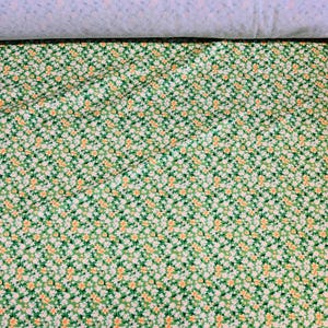 May include: Green fabric featuring a repeating pattern of small white and yellow flowers. The fabric is quilted, with a light blue and white striped fabric visible at the top.
