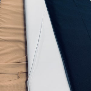 Könnte beinhalten: Nahaufnahme von drei Stoffrollen in verschiedenen Farben: Beige, Weiß und Marineblau. Der Stoff ist eng gerollt, was auf ein glattes Material hindeutet. Das Bild ist gut beleuchtet und zeigt die Textur und Farbvariationen der Rollen.