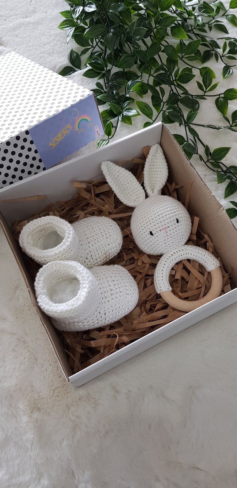May include: A white gift box filled with brown packing paper. The box contains a pair of white crocheted baby booties, a white crocheted bunny rattle, and a wooden teething ring. The box has a label that says "Sisters Baby Shop".
