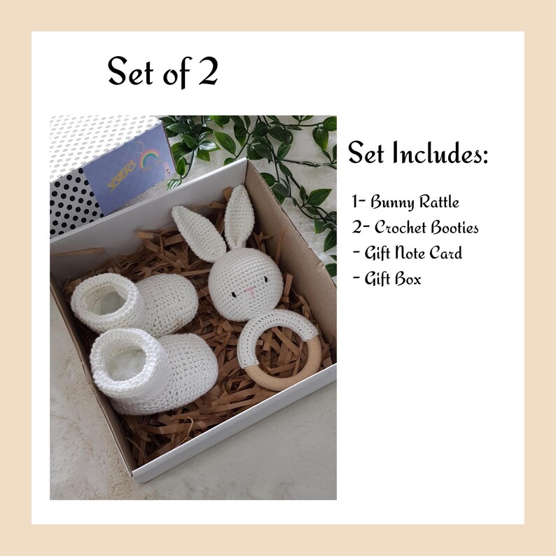 May include: A white crocheted bunny rattle, two white crocheted booties, a wooden ring, and a gift note card are displayed in a white gift box with brown shredded paper. The box is labeled "Set Includes: 1- Bunny Rattle, 2- Crochet Booties, Gift Note Card, Gift Box".