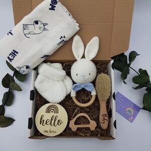 May include: A gift box filled with baby items, including a white crocheted bunny, a wooden rattle, a wooden baby brush, a white crocheted mitten, a wooden car, and a wooden sign that says "hello i'm here".