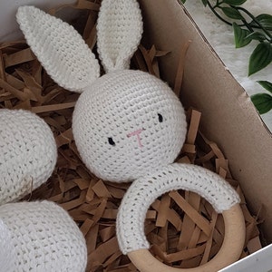May include: A white crocheted bunny rattle with a wooden ring. The bunny has black eyes and a pink nose. The rattle is in a white gift box with brown paper shreds.