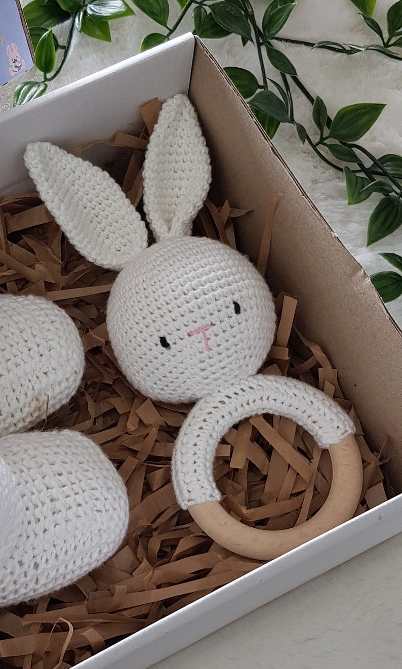 May include: A white crocheted bunny rattle with a wooden ring. The bunny has black eyes and a pink nose. The rattle is in a white gift box with brown paper shreds.
