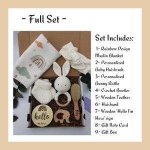 May include: A gift box filled with baby items, including a rainbow muslin blanket, a crocheted bunny rattle, a wooden teether, a hairband, a wooden sign that says "hello I'm here", a crochet bootie, a baby hairbrush, and a gift note card. The box is decorated with black and white polka dots.