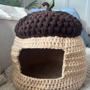 May include: A handmade crochet pet bed shaped like an acorn. The top is dark brown, with a green stem and leaf, and the base is tan. The bed has a rectangular opening for easy access for a small pet. The bed is made of soft yarn.