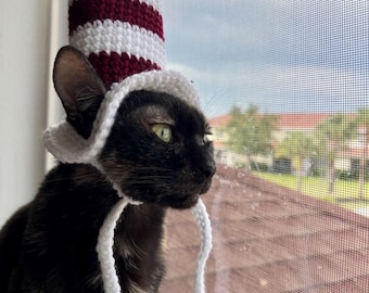 May include: A tall, cylindrical crochet hat featuring alternating horizontal stripes of deep maroon and white. The hat includes a white brim and two long, thin white crochet ties that hang down from the sides, worn by a dark-furred cat.