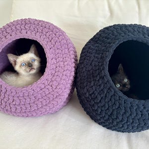 May include: Two round, chunky knit pet beds, one in lavender and one in black, each featuring a circular opening. A light-colored kitten with blue eyes sits inside the lavender bed, while a black kitten rests inside the black bed.