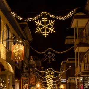 May include: A narrow street lined with buildings decorated with twinkling lights and snowflakes. The street is covered in snow and there are Christmas trees in front of the buildings.