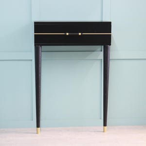 May include: A black console table with a single drawer, featuring a gold accent line and handle. The table has four slender, dark legs with gold-colored tips. The background is a light blue wall.