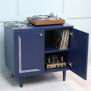 Op de afbeelding: Een marineblauwe platenkast met een houten platenspeler erop. De kast heeft zilveren accenten en een deur die openstaat om vinylplaten en audioapparatuur te onthullen. De kast staat op korte, afgeronde poten.