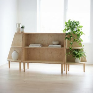 Op de afbeelding: Een lichtgekleurde houten kast met planken en een gebogen basis. De kast heeft een uniek ontwerp met schuine zijkanten en wordt ondersteund door vier houten poten. Decoratieve items, waaronder planten en vazen, worden op de planken weergegeven.