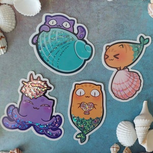 May include: Four colourful stickers featuring cartoon cats dressed as mermaids. The stickers are on a light blue background. The cats are wearing seashells, holding hearts, and have sparkly tails.
