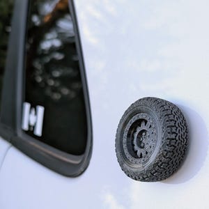 May include: A black tire with a detailed tread pattern and a black rim is attached to a white car. The tire is a small, decorative model.