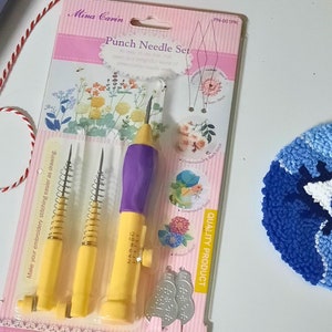 May include: A set of three punch needles with yellow handles and purple tips, packaged on a pink and white card with the text "Mina Carin Punch Needle Set" and "PN-001PK". The needles are used for embroidery and are shown next to a small blue and white eye-shaped rug.