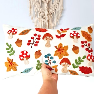 May include: White rectangular cushion with a colourful embroidered design of mushrooms, leaves, acorns, and berries.