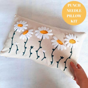 Op de afbeelding: Een wit kussen met geborduurde witte madeliefjes met gele centra en groene stelen. Het kussen is gelabeld "Punch Needle Pillow Kit".