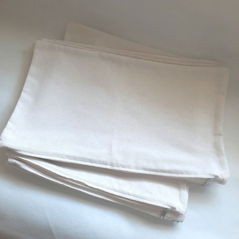 May include: A stack of three white canvas pillowcases with zippers. The pillowcases are folded and stacked on top of each other.
