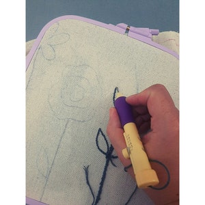 May include: A close-up of a hand holding a purple and yellow embroidery tool with a needle and thread. The tool is being used to stitch a design on a white fabric stretched in an embroidery hoop. The design is a simple flower with a stem and leaves.