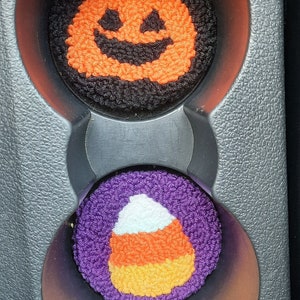 May include: Two Halloween-themed car cup holders. One is a black and orange pumpkin with a smiling face. The other is a purple candy corn with white and orange stripes.