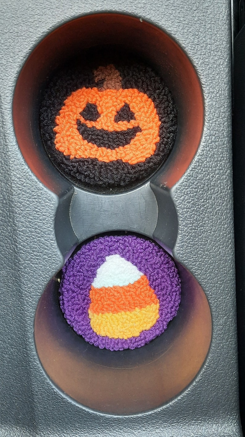 May include: Two Halloween-themed car cup holders. One is a black and orange pumpkin with a smiling face. The other is a purple candy corn with white and orange stripes.