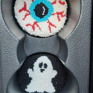 May include: Two crocheted car cup holders, one with a white eyeball with red veins and a blue iris, and the other with a white ghost on a black background.