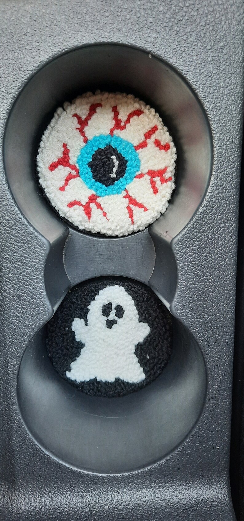 May include: Two crocheted car cup holders, one with a white eyeball with red veins and a blue iris, and the other with a white ghost on a black background.