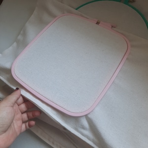 May include: A pink embroidery hoop with a white fabric stretched across the frame. The hoop is lying on a white fabric surface.