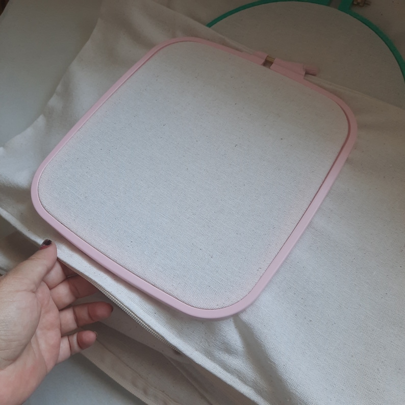 May include: A pink embroidery hoop with a white fabric stretched across the frame. The hoop is lying on a white fabric surface.