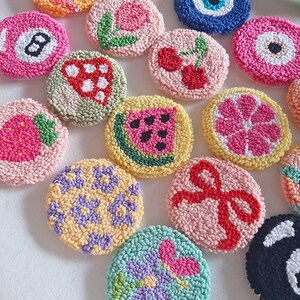 May include: A collection of colorful, round, handmade coasters with various designs. The coasters feature images of a watermelon slice, cherries, a strawberry, a flower, a bow, and an eight ball. The coasters are made with a textured, looped yarn.