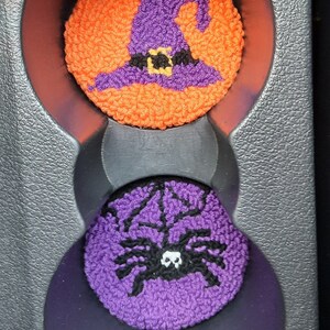 May include: Two Halloween-themed car cup holders. The top one is orange with a purple witch's hat and a black belt. The bottom one is purple with a black spider.
