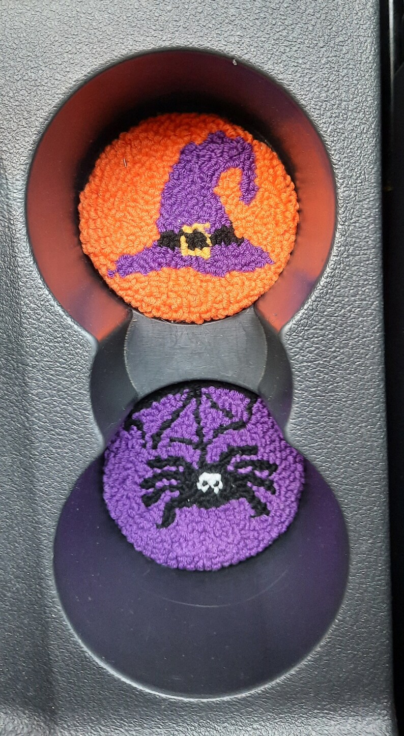 May include: Two Halloween-themed car cup holders. The top one is orange with a purple witch's hat and a black belt. The bottom one is purple with a black spider.