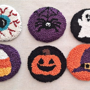 May include: Six handmade Halloween-themed coasters. The coasters are round and feature various designs, including an eye, a spider, a ghost, a candy corn, a pumpkin, and a witch's hat. The coasters are made with a variety of colours, including white, red, blue, purple, orange, black, and yellow.