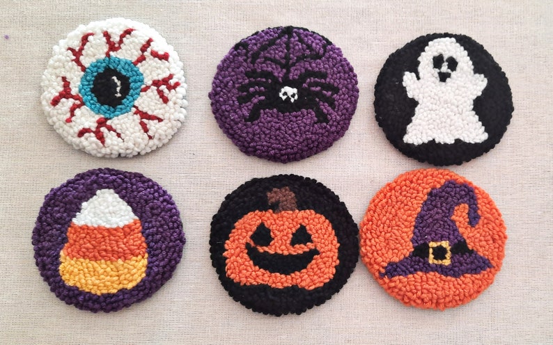 May include: Six handmade Halloween-themed coasters. The coasters are round and feature various designs, including an eye, a spider, a ghost, a candy corn, a pumpkin, and a witch's hat. The coasters are made with a variety of colours, including white, red, blue, purple, orange, black, and yellow.