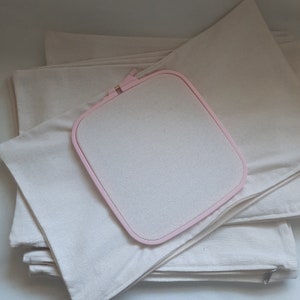 May include: A stack of white fabric squares with a pink embroidery hoop on top. The fabric squares have zippers on the sides.