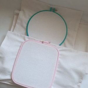 May include: Two embroidery hoops, one pink and one green, are on top of a white fabric. The hoops are empty and ready for embroidery.