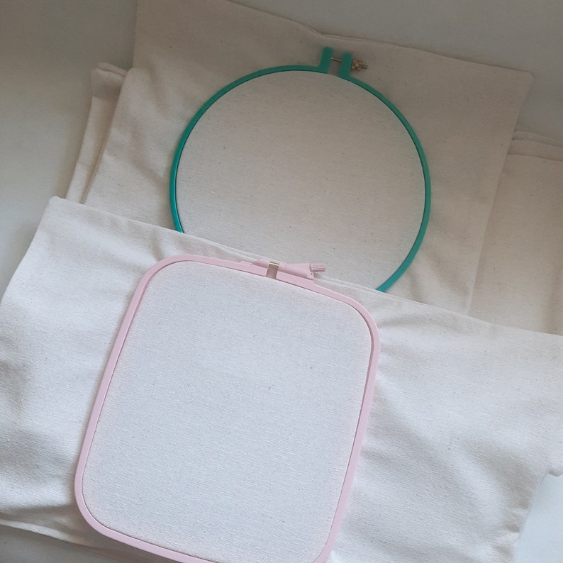 May include: Two embroidery hoops, one pink and one green, are on top of a white fabric. The hoops are empty and ready for embroidery.